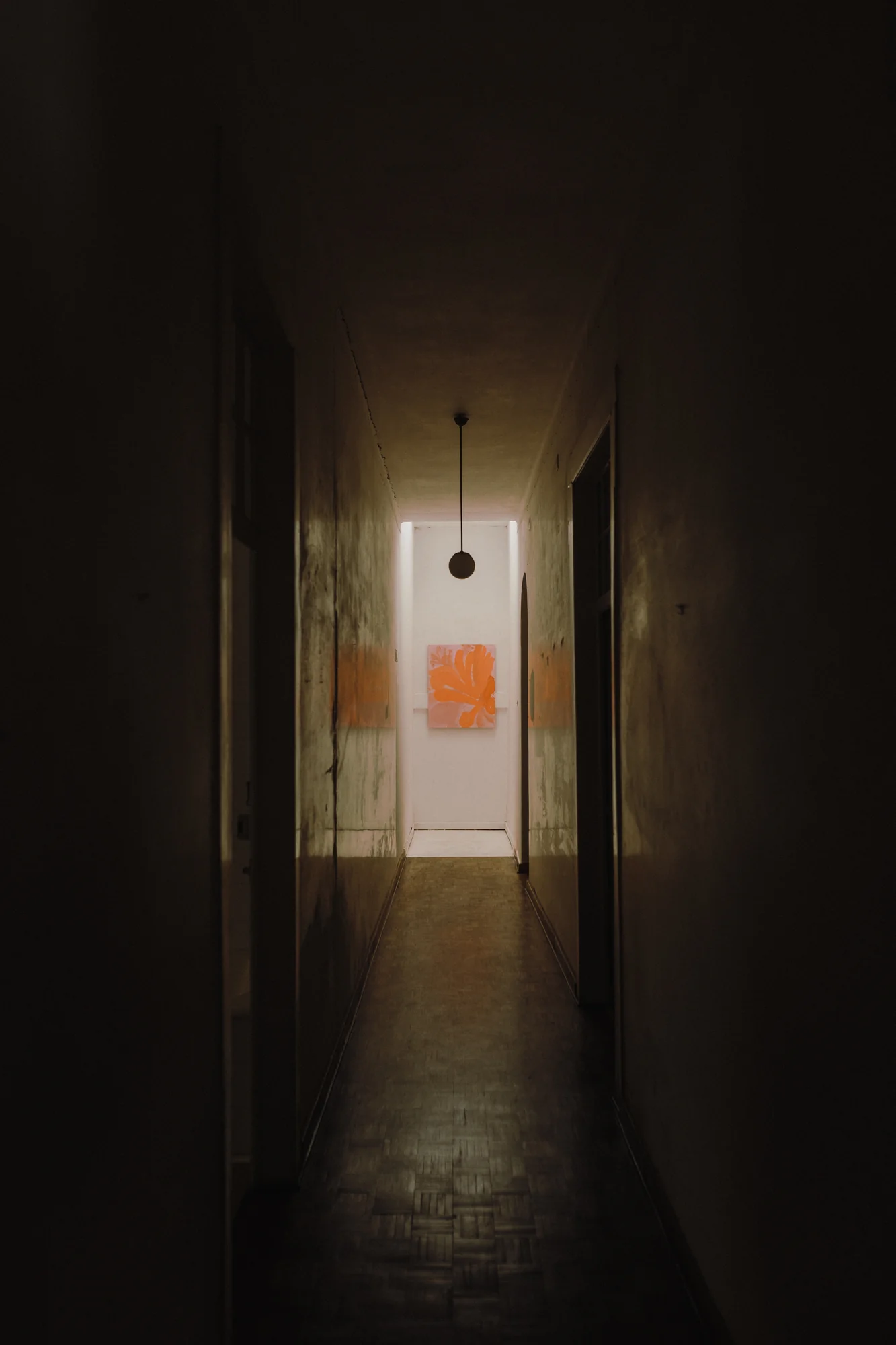 Corredor escuro que termina numa porta iluminada com uma pintura floral laranja