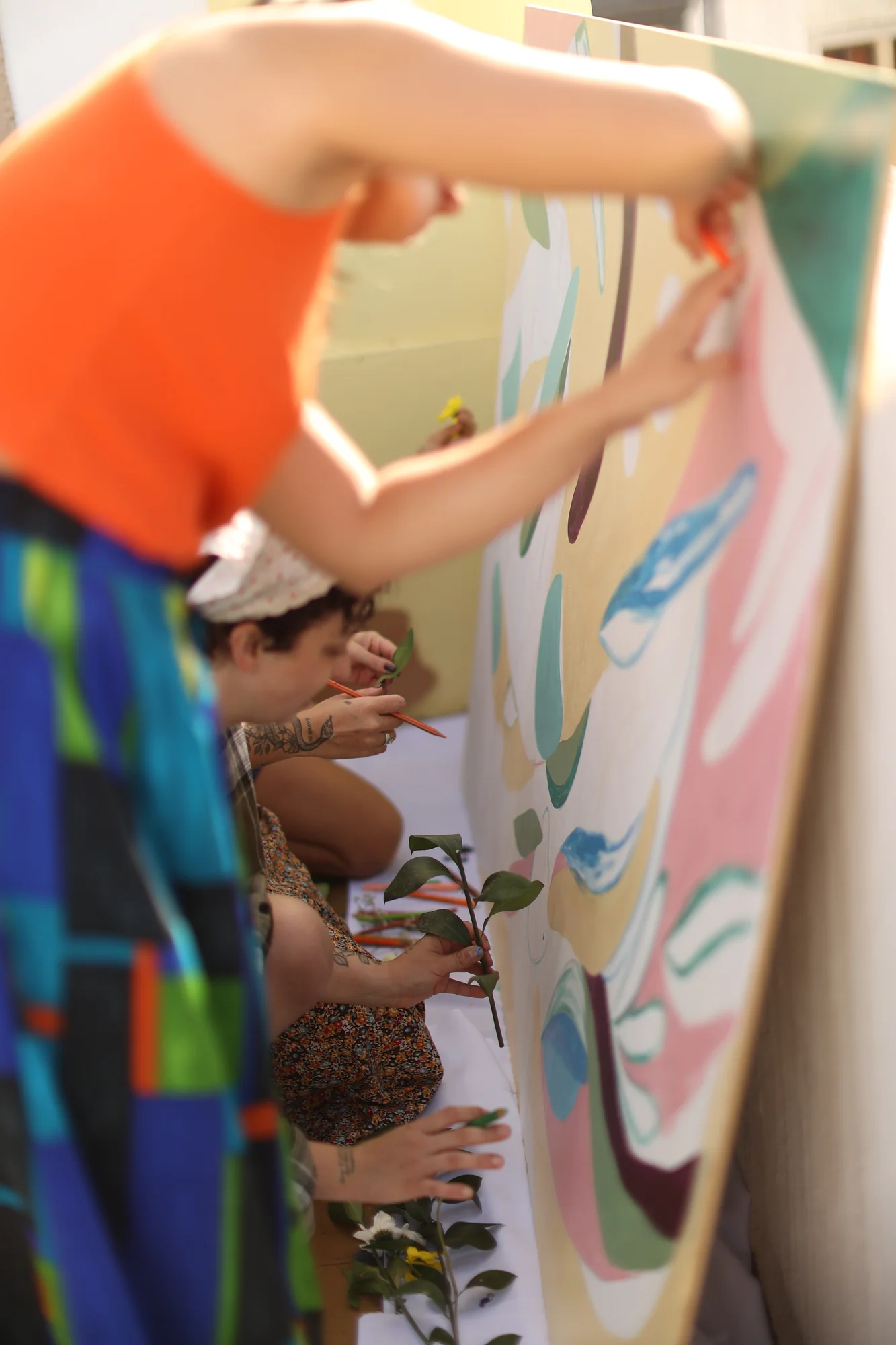 Duas pessoas pintam um painel grande ao ar livre, uma de pé com pincel e outra agachada.